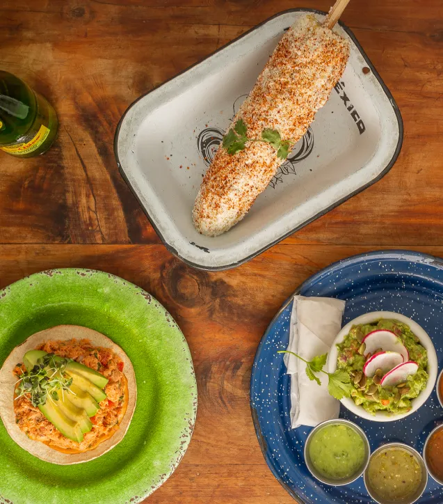 Elote dusted with chile on an enamel tray, next to plates of guacamole and salsa on a wooden table